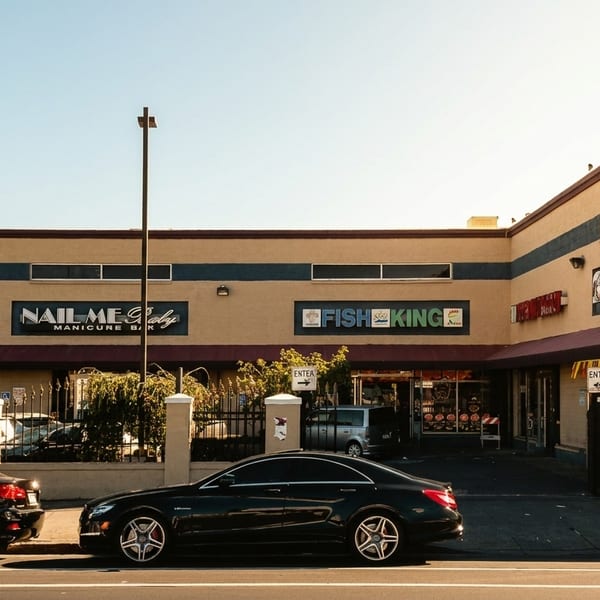 Exterior of a strip mall with Little Caesars, Fish King, and Nail Me Baby businesses, with cars parked.